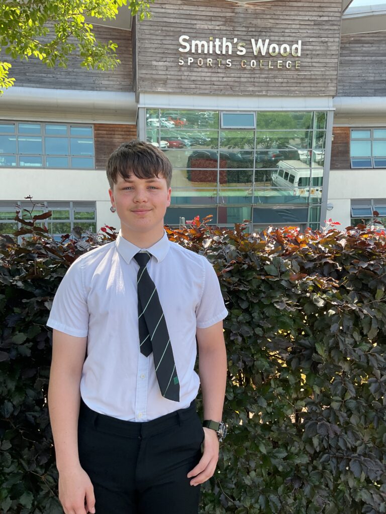 Student in school uniform standing outdoors in front of the Smith’s Wood Sports College building, with greenery in the foreground and windows reflecting parked cars behind