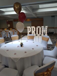 Round tables set for a prom event with white tablecloths, gold chair sashes, and small centrepieces, alongside red, white, and gold helium balloons. In the background, large illuminated letters spell “PROM” next to a light-up dance floor in a decorated function room