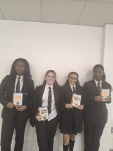 Four students in school uniform stand against a white brick wall, each holding a copy of the book Think Twice by Alex Bellos. The students are posed side by side for a photograph in a bright indoor school setting.