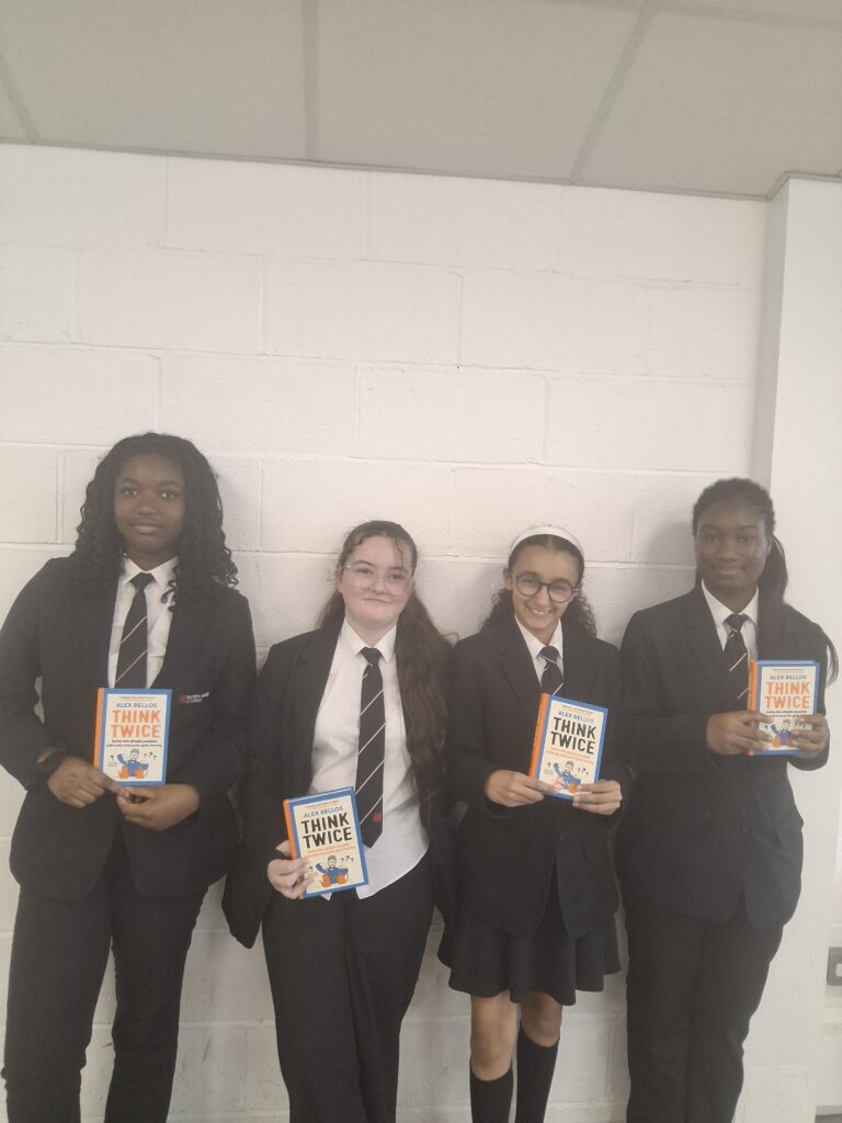 Four students in school uniform stand against a white brick wall, each holding a copy of the book Think Twice by Alex Bellos. The students are posed side by side for a photograph in a bright indoor school setting.
