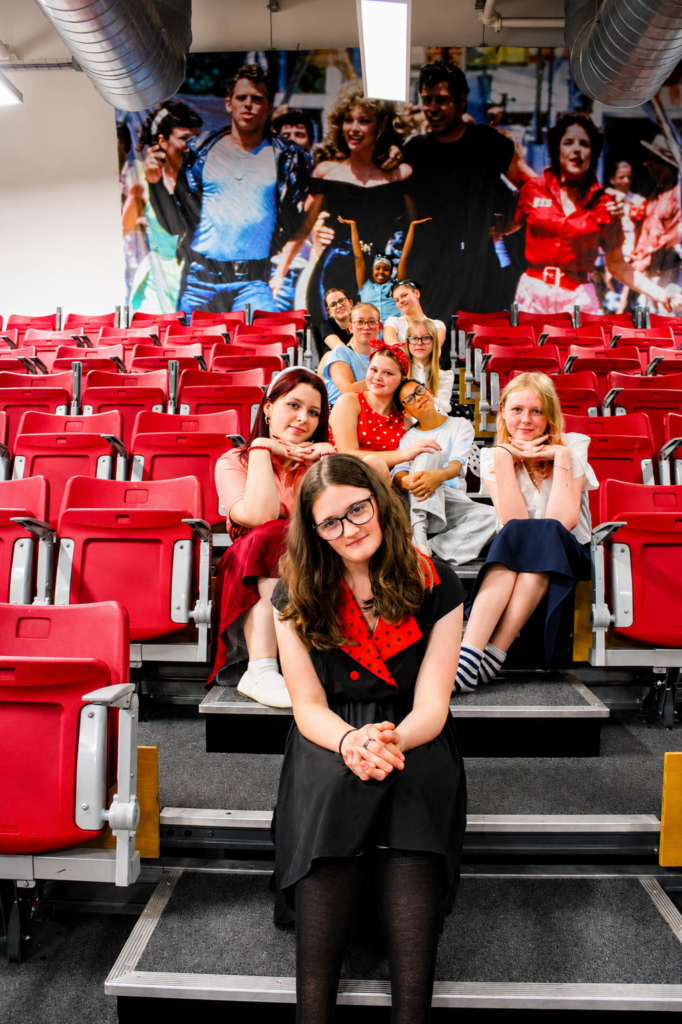 Students from the Smith’s Wood Academy production of Grease sit on red tiered theatre seating in costume for a cast photograph. A large mural inspired by Grease is displayed behind them, with the cast arranged in rows up the seating, creating a staged promotional image ahead of the school production.