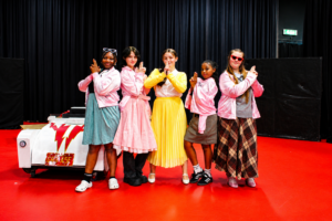 Five students in 1950s-inspired costumes pose on a stage during rehearsals for Smith’s Wood Academy’s production of Grease. The group stands beside a prop car, wearing pink jackets and colourful period-style outfits against a backdrop of black stage curtains and a bright red floor