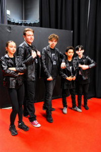 Five students in black leather jackets and dark clothing pose together on a stage with black curtains and a red floor during rehearsals for Smith’s Wood Academy’s production of Grease. The group is standing in character in front of a stage set, reflecting the show's iconic 1950s greaser style.