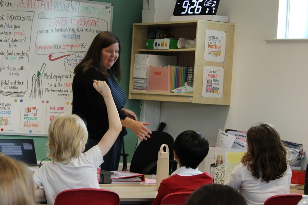 A classroom scene during a Year 5 workshop. Several pupils sit around a table while one pupil raises a hand to answer a question. An adult stands at the front of the room leading the discussion. In the background, classroom displays show homework information, school expectations and learning posters, with shelves of books and resources above a workstation.