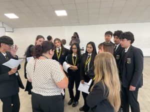 A group of students in school uniform stand in a circle around an adult facilitator in a large hall, listening to instructions and holding sheets of paper during a collaborative learning activity. The room has white walls, a light wood floor, and bright overhead lighting.